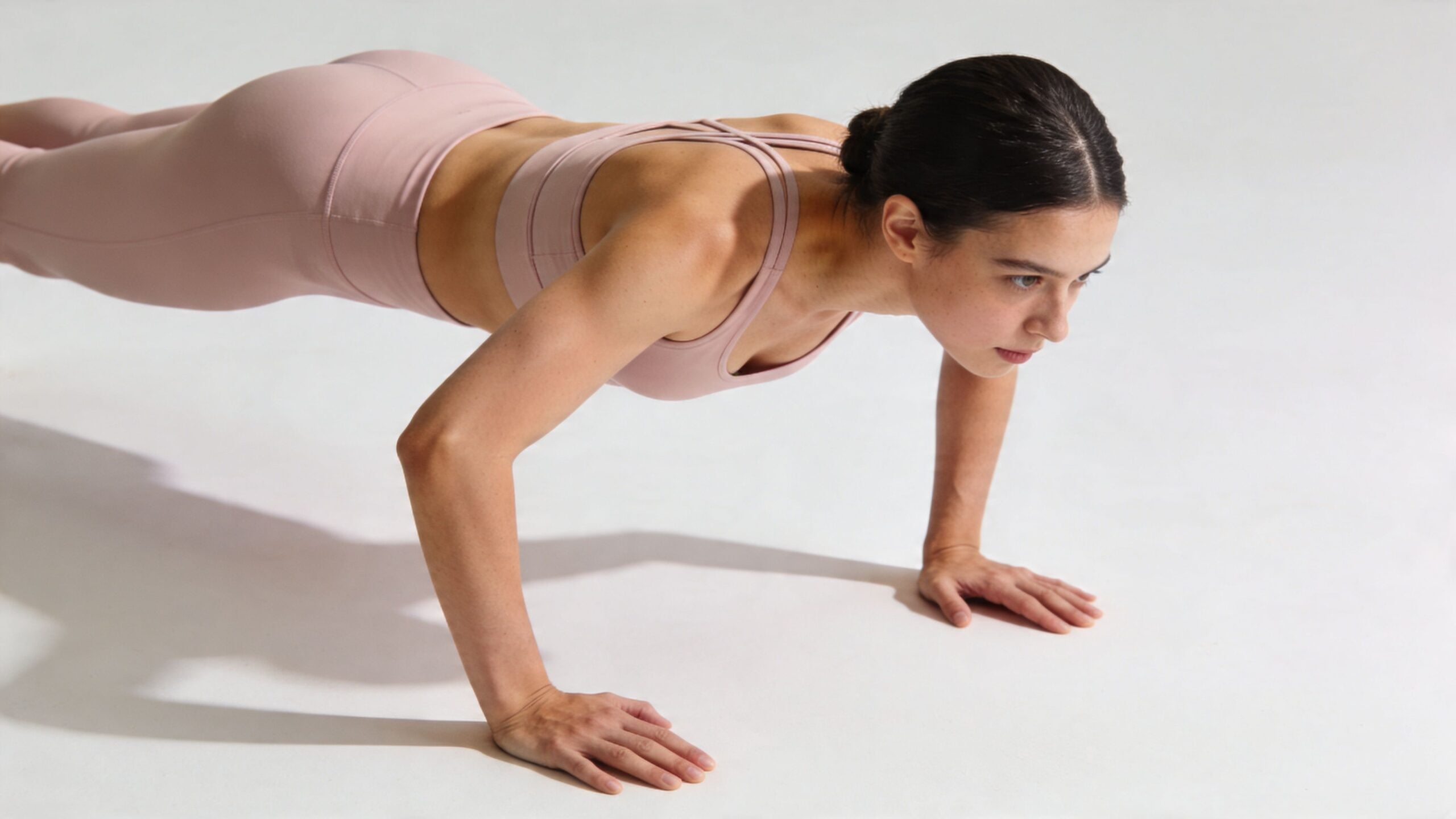 A fit young woman performing a push-up on a plain white floor in her activewear clothing.