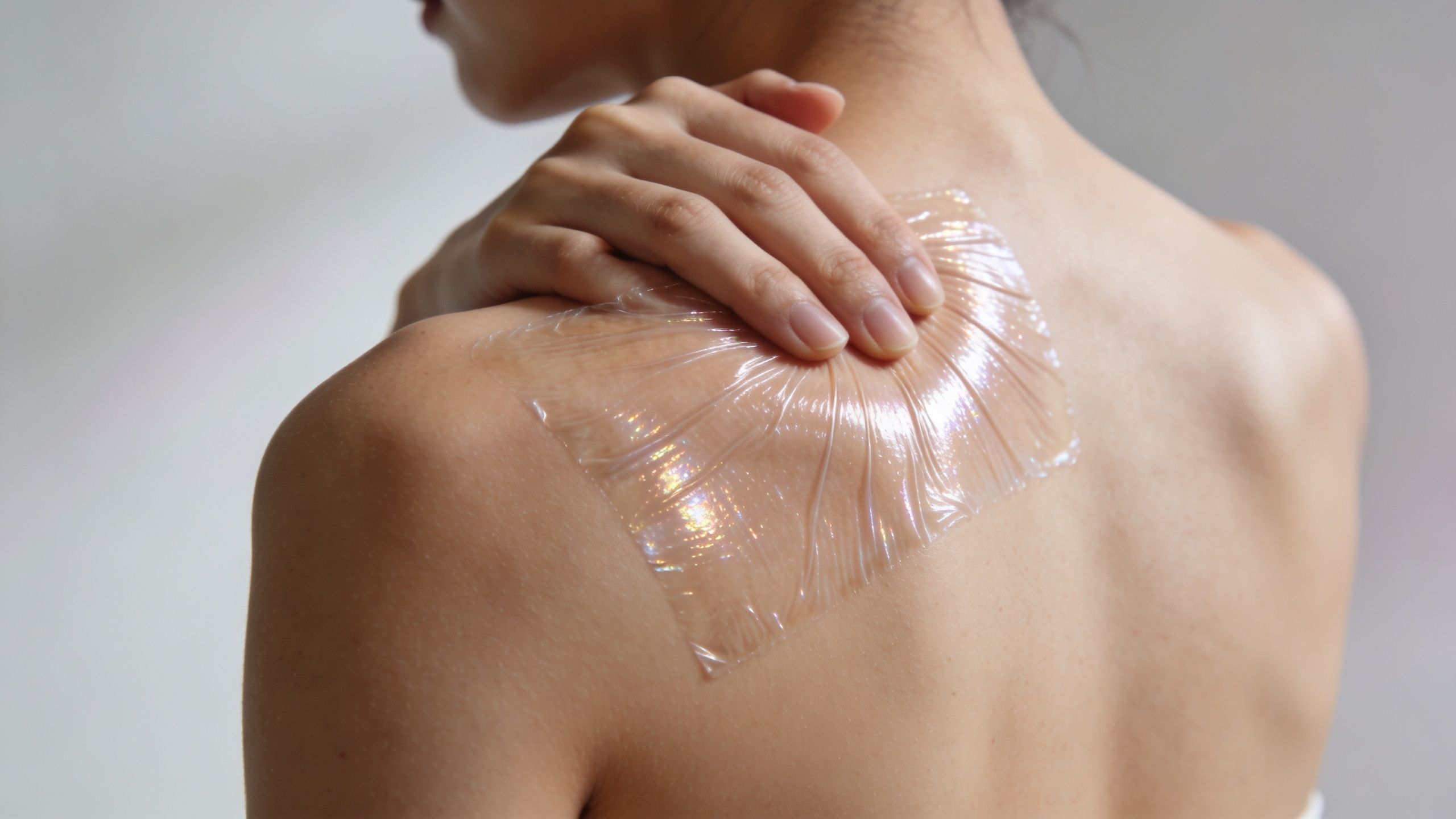 A person applying a clear adhesive medical patch to their shoulder for back pain relief therapy.
