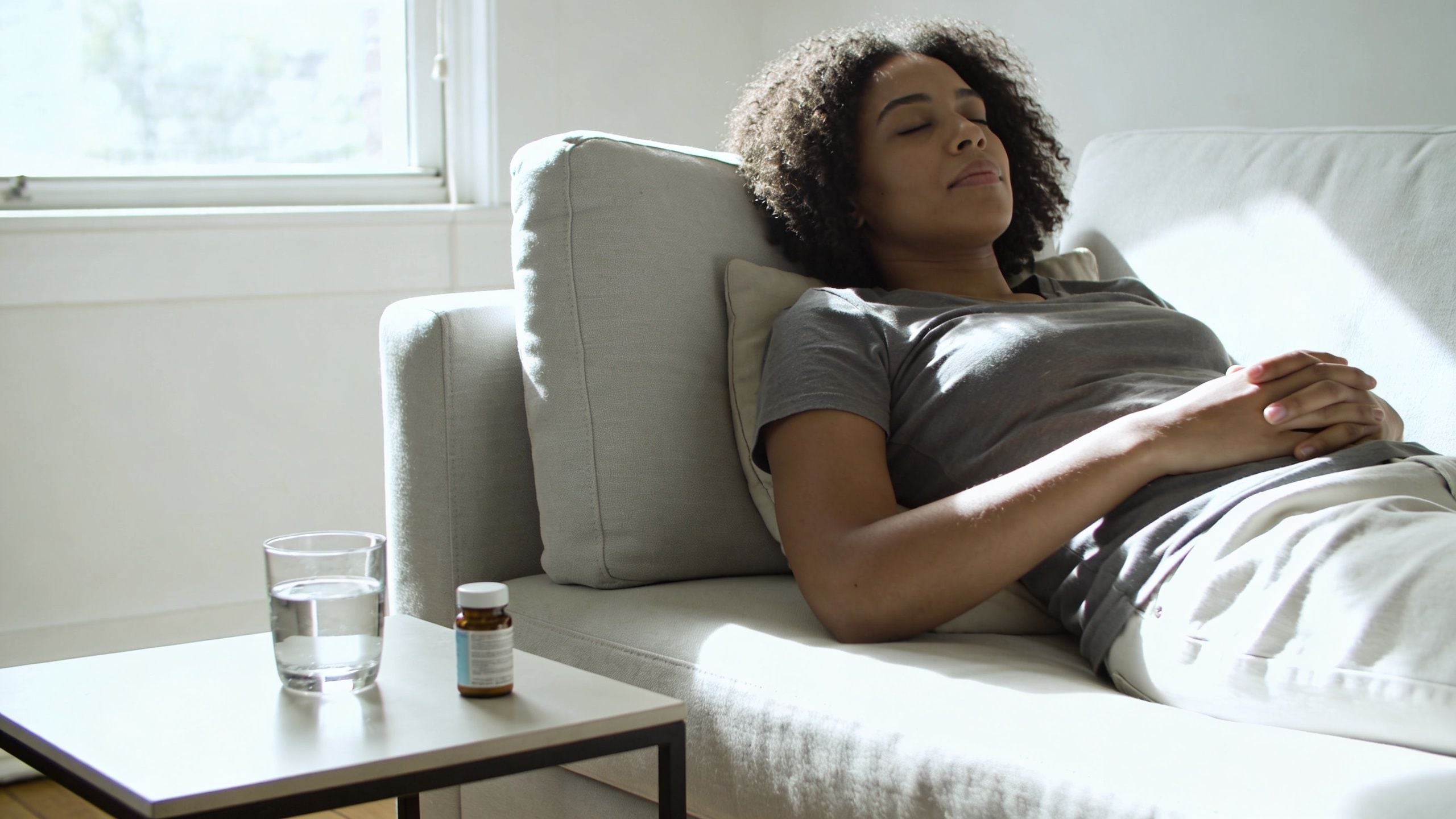 A woman resting on a comfortable sofa with a glass of water and medication nearby for recovery.